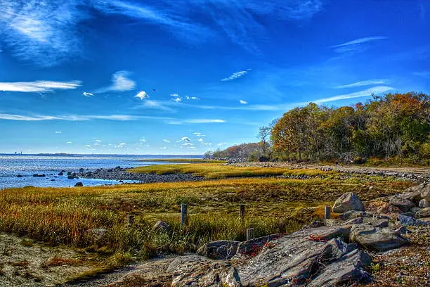 A DSLR photo taken in Greenwich Point Park in autumn. It is a clear, crisp day, with wisps of clouds, showing an HDR image of a shoreline pathway meandering along the ocean, with trees covered in colorful autumn foliage in the background. The photo was taken in Greenwich, Connecticut.
