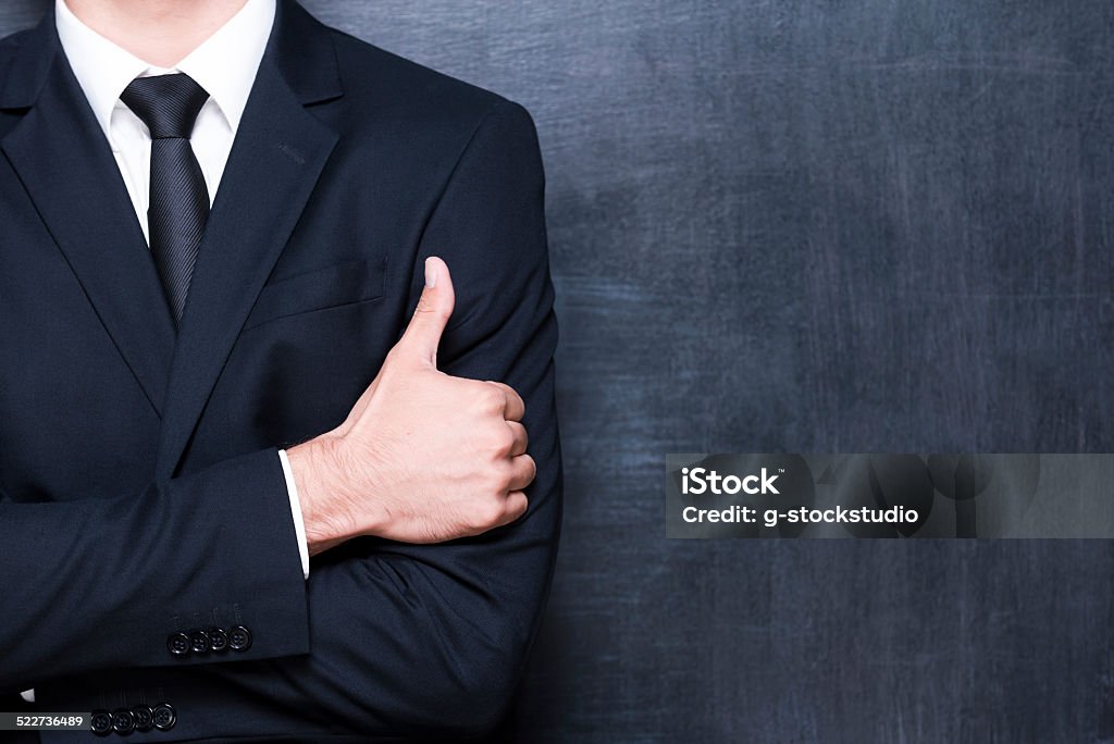 Excellent! Close-up of man in formalwear showing his thumb up while standing against blackboard Achievement Stock Photo
