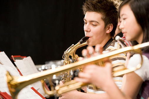 Saxophone player standing near music stand with notes and playing saxophone while having concert rehearsal with musicians under blue sky