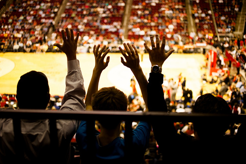 Unrecognizable people raise hands in excitement during a basketball sports game inside a large basketball sports stadium. Basketball court seen below. Silhouette. Cheering fans.