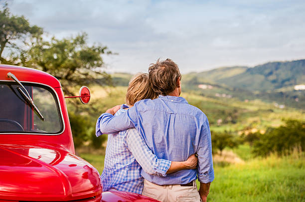 Senior couple hugging, vintage styled red car, sunny nature Senior couple hugging, vintage styled red car, green sunny nature, rear view the farmer and his wife pictures stock pictures, royalty-free photos & images