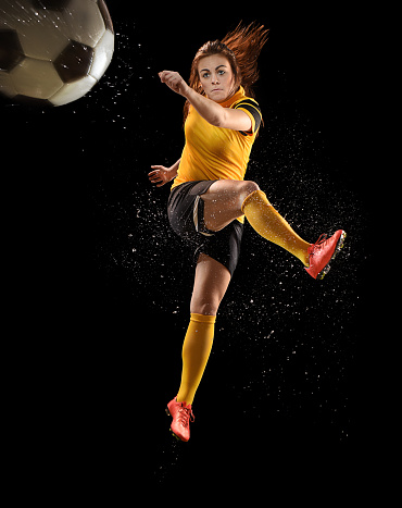 young woman football leaps into the air and smashes the ball to camera position.the background is pure black and water splashes can be seen as a result of the impact of foot on ball . She is wearing a gold kit and orange football boots.