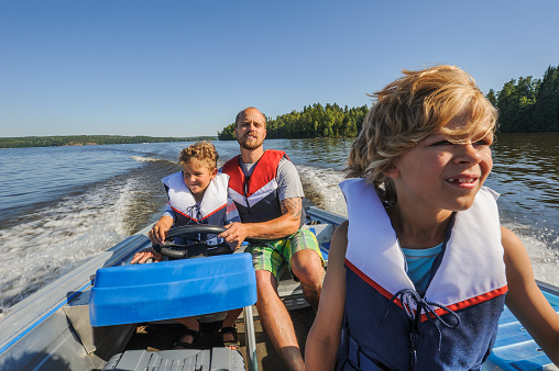 Father and sons out boating together. The father lets one of the boys steer the boat. They're wearing life vests. In the background there's forested shore line, water, and blue sky.