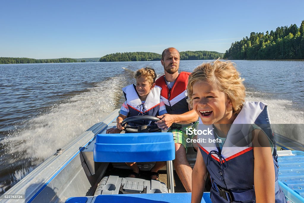 father and sons boating Father and sons out boating together. The father lets one of the boys steer the boat. They're wearing life vests. In the background there's forested shore line, water, and blue sky. Nautical Vessel Stock Photo