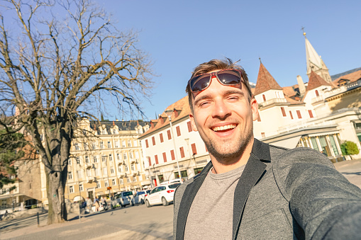 Young handsome man taking selfie at Meran old town in South Tyrol in Italy - Adventure and travel lifestyle around european destination - Composition with tilted horizon and warm afternoon color tones