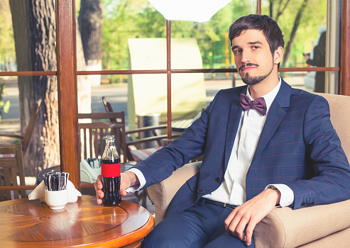 Handsome male sitting at cafe and holding a fresh drink cola. Businessman dressed in fashion costume. Stylish man