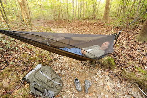 Man resting or sleeping in camping hammock on hiking trail in woods with backpack and shoes nearby.  Listening to mp3 or ipod.