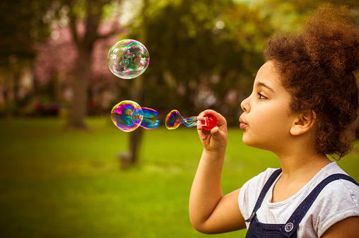 Stock photo of a little girl blowing soap bubbles in playground. Profile shot and natural light.  Shot in Raw and post processed in ProPhoto RGB. No sharpening applied. This file has a 