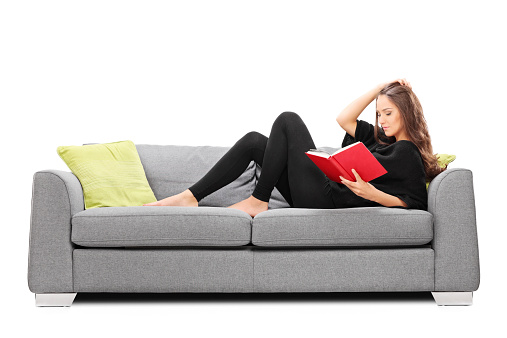 Relaxed young woman reading a book seated on sofa isolated on white background