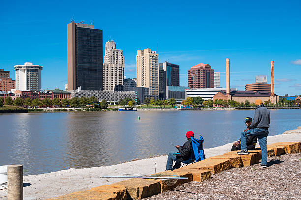 A tranquil view of Cleveland Ohio's urban skyline with a white lighthouse on Lake Erie, captured on a sunny day. The scene highlights the city's modern architecture, serene water, and vibrant cityscape.