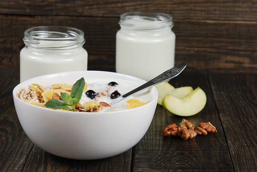 Oat, wheat and corn flakes with nuts and yogurt in small white bowls with a spoon and apples on a dark wooden background