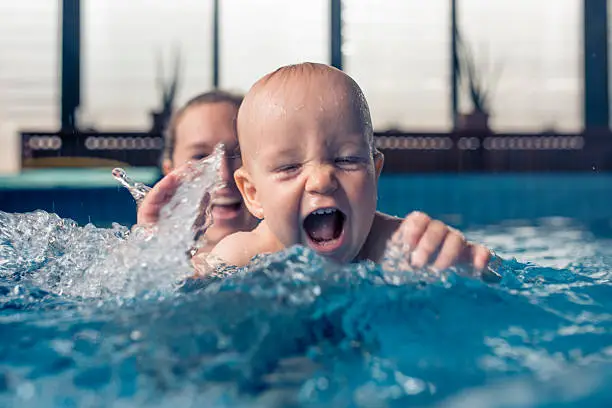 Nine month old baby boy at his first swimming lesson. Looking at camera, smiling and enjoying water. Mother is holding him and helping him to swim.