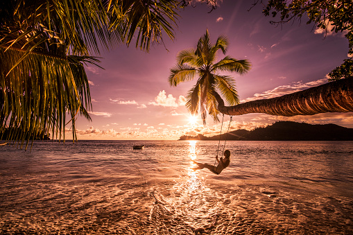 Young happy woman enjoying in beautiful sunset on the beach while swinging above the water.
