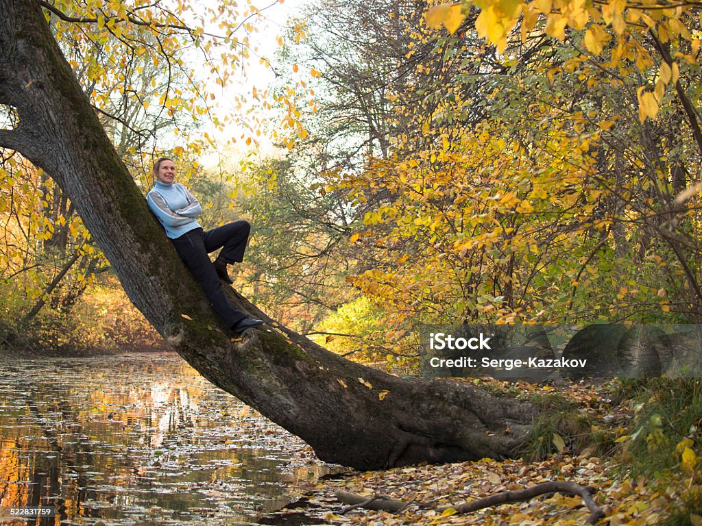 Young woman stands on a tree and smiling A young woman stands on a tree and smiling on a background of yellow leaves Adult Stock Photo