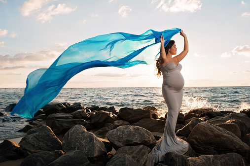 A beautiful, smiling pregnant woman on a rocky beach at sunrise. She is holding a blue silk veil that's flowing in the wind.