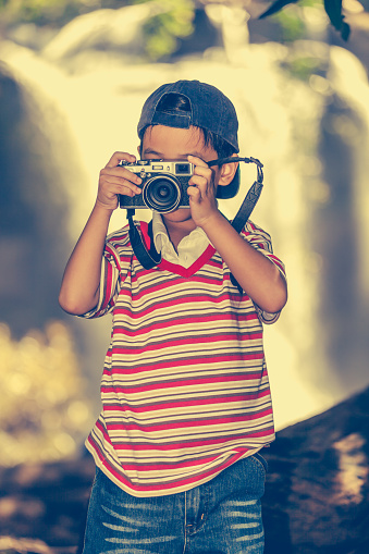 Asian child taking photo by professional digital camera on blurred waterfall background. Handsome boy in nature. Outdoors portrait. Vintage picture style.