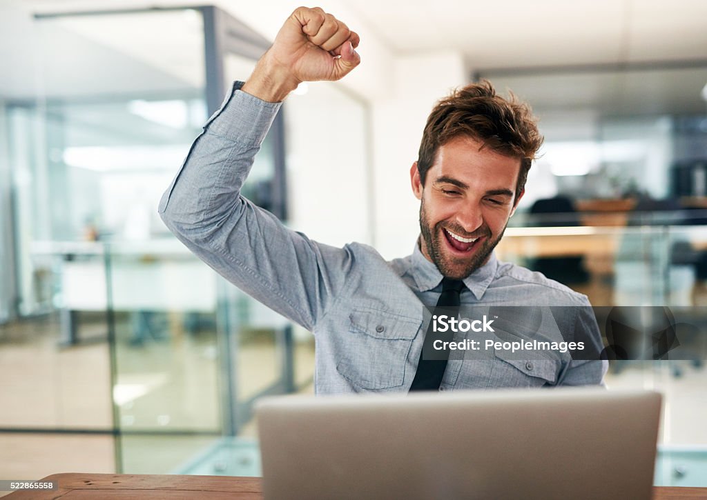 Yes to success! Shot of a young businessman cheering while using a laptop at his desk in an office Yes - Single Word Stock Photo