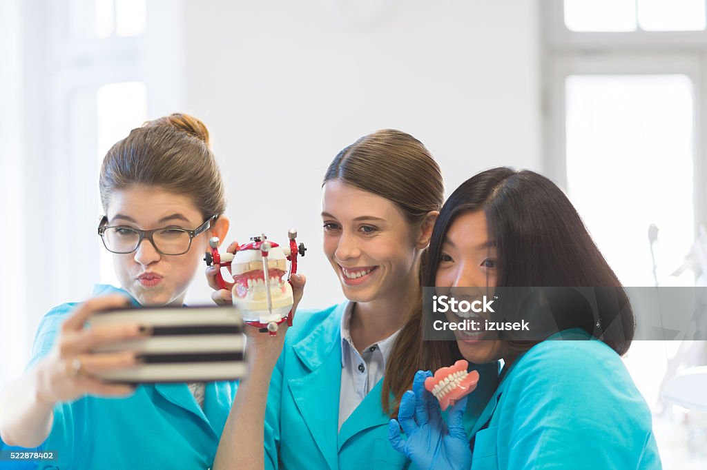 Playful female students learning prosthetic dentistry, taking selfie Playful multi ethnic female students in a prosthodontic lab, holding dentures and taking selfie using smart phone. Dentist Stock Photo
