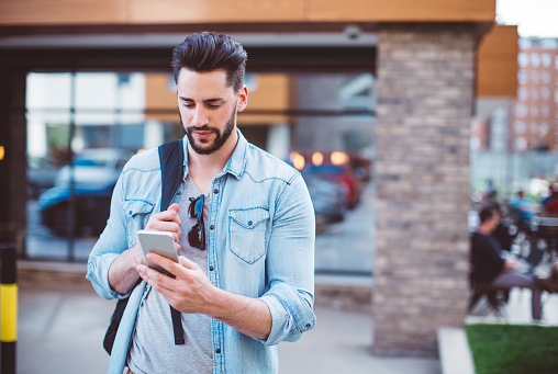 Young urban professional man using smart phone. Carry backpack on one shoulder and sunglasses atached on his t-shirt.