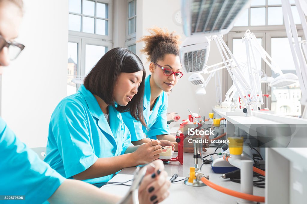 Multi ethnic female students learning prosthetic dentistry Multi ethnic female students - asian and afro american - wearing uniforms in a prosthodontic lab, learning prosthetic dentistry. Dentist Stock Photo