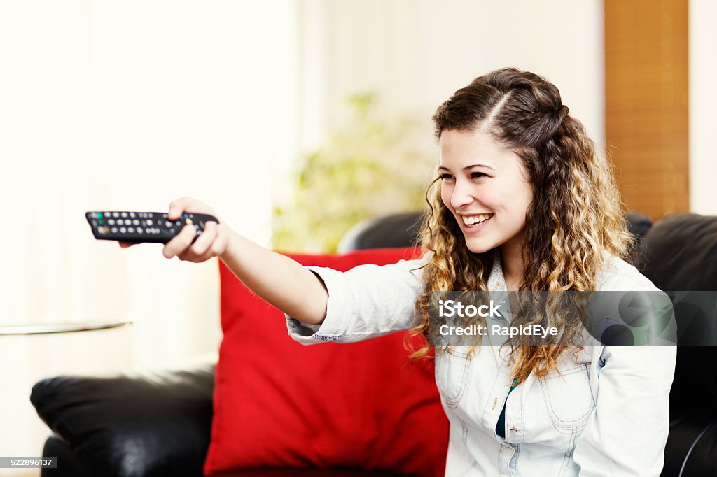 Smiling young beauty selecting TV program with remote Beautiful, smiling young woman using TV remote control. Copy space on the window behind her. Adult Stock Photo
