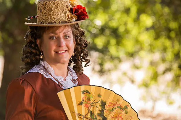 A beautiful woman dressed in the style of the 1880's in California holding a fan with a beautiful soft Boke background.