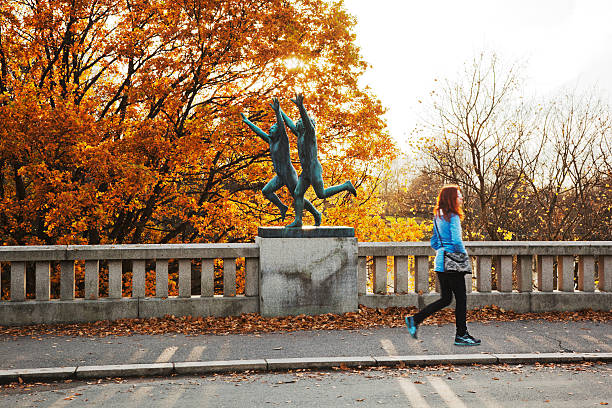 Oslo, Norway - October 30, 2014: Orange oak tree  and woman with red hair passing a sculpture in Gustav Vigeland Sculpture park at sunset in fall.