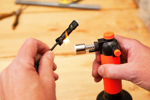 Male hands are using a butane torch on a heat shrink tubing electrical connection. The tubing will encase a connection, in this case a Micro USB adapter. The background is a maple work bench with needle-nose pliers and a portion of a square. The thumbnails are dirty when viewed at 100%.