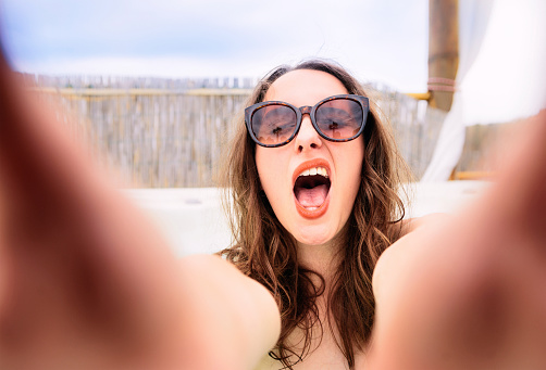 A young woman making a funny face as she holds a smartphone to take a selfie on vacation.