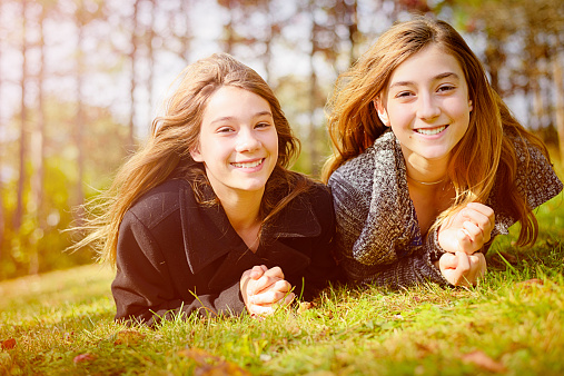 Two happy smiling pre-teen sisters in warm clothing, lying together on their front in a grassy meadow, looking at camera.