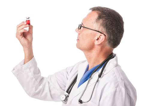 Side view of a mature caucasian male doctor or scientist holding up and examining a vial containing a red liquid. He is wearing a white lab coat and is isolated on a pure white background.