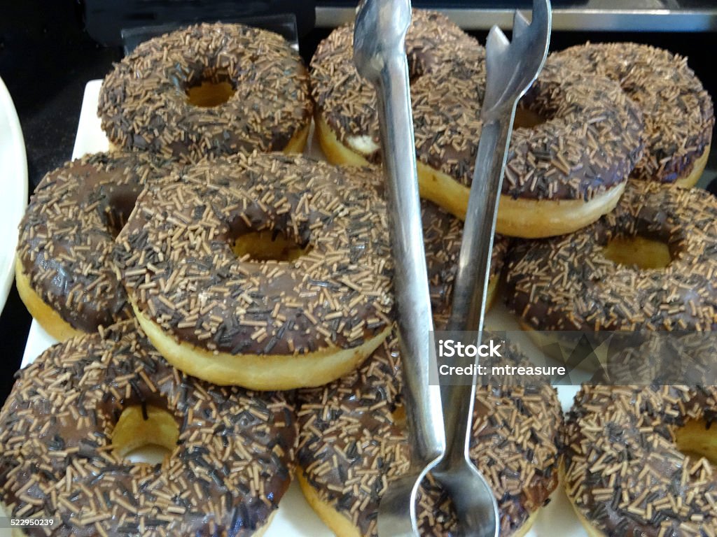 Image of freshly cooked ring doughnuts with chocolate icing / sprinkles Photo showing freshly cooked ring doughnuts that have been decorated with chocolate icing and sprinkles.  The doughnuts are displayed on a white plate with stainless steel kitchen tongs, so that you are able to 'serve yourself'. Baked Stock Photo