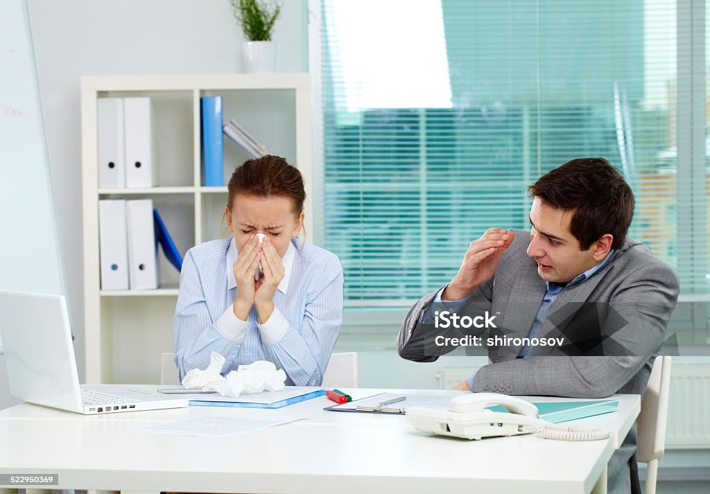 Sick in office Image of businesswoman sneezing while her partner looking at her unsurely in office People Stock Photo