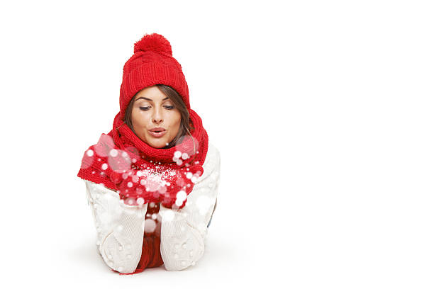 Winter, christmas, holidays concept. Beautiful woman in red winter hat, scarf and mittens lying down and blowing snow flakes at camera, looking at palms, over white background