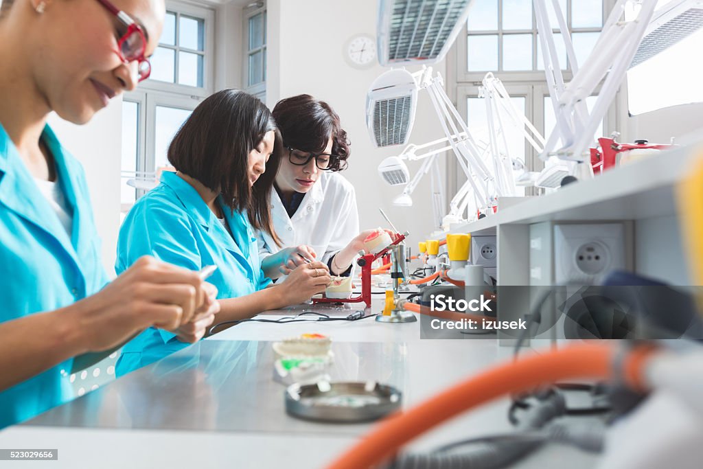 Multi ethnic female students learning prosthetic dentistry Multi ethnic female students - asian and afro american - wearing uniforms in a prosthodontic lab, learning prosthetic dentistry. Asian woman talking with teacher. Dental Health Stock Photo