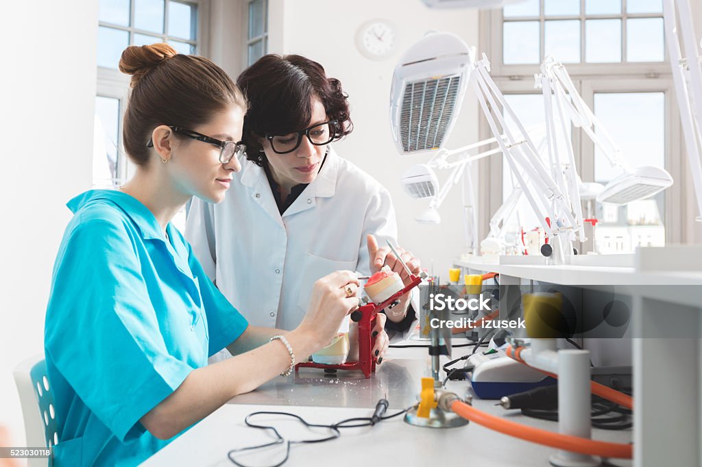 Young woman learning prosthetic dentistry, talking with teacher Female student wearing uniform sitting in a prosthodontic lab, learning prosthetic dentistry, talking with her teacher. Adult Stock Photo
