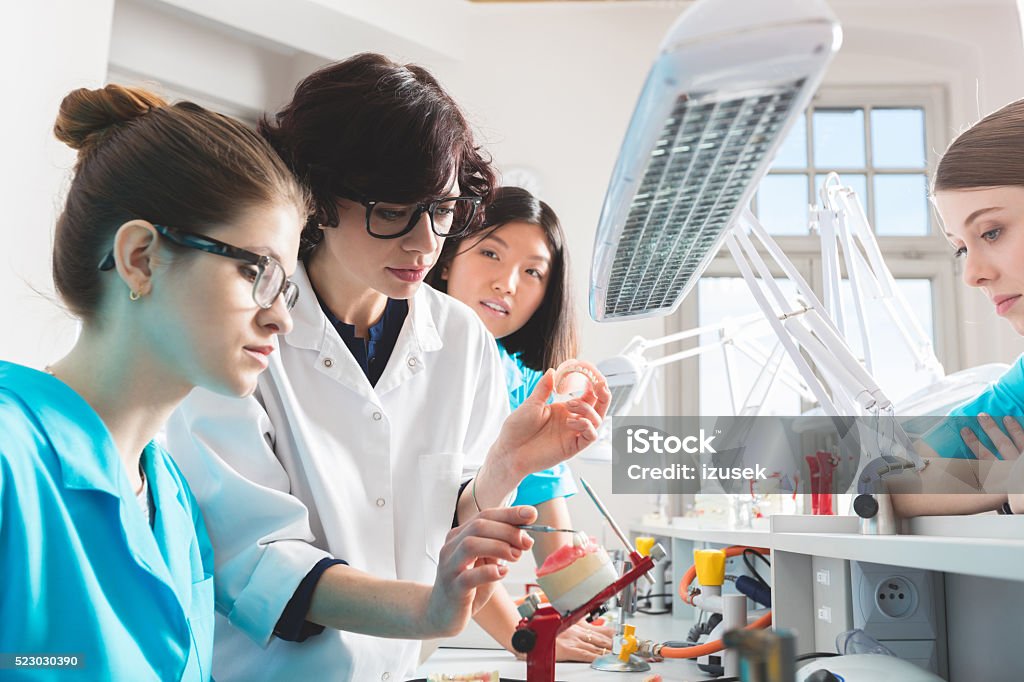 Female students learning prosthetic dentistry, talking with teacher Multi ethnic female students wearing uniform sitting in a prosthodontic lab, learning prosthetic dentistry, talking with her teacher. Adult Stock Photo