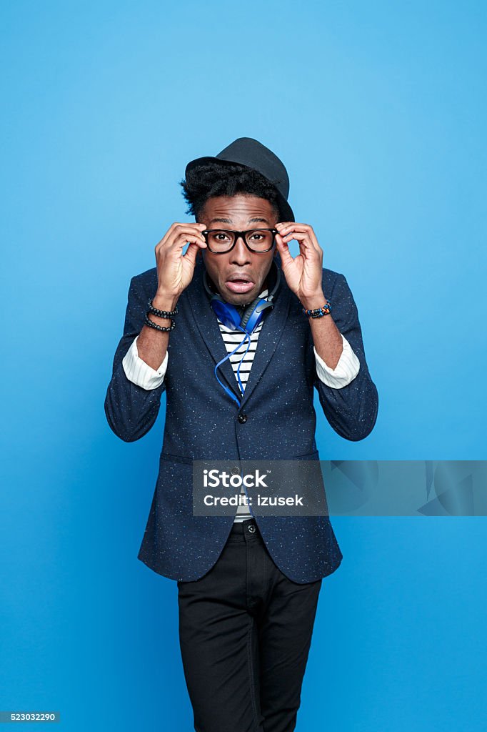 Crazy afro american guy in fashionable outfit Studio portrait of crazy, surprised afro american young man wearing striped top, navy blue jacket, nerd glasses, hat and headphone, looking at camera, making funny face. Studio portrait, blue background. Men Stock Photo