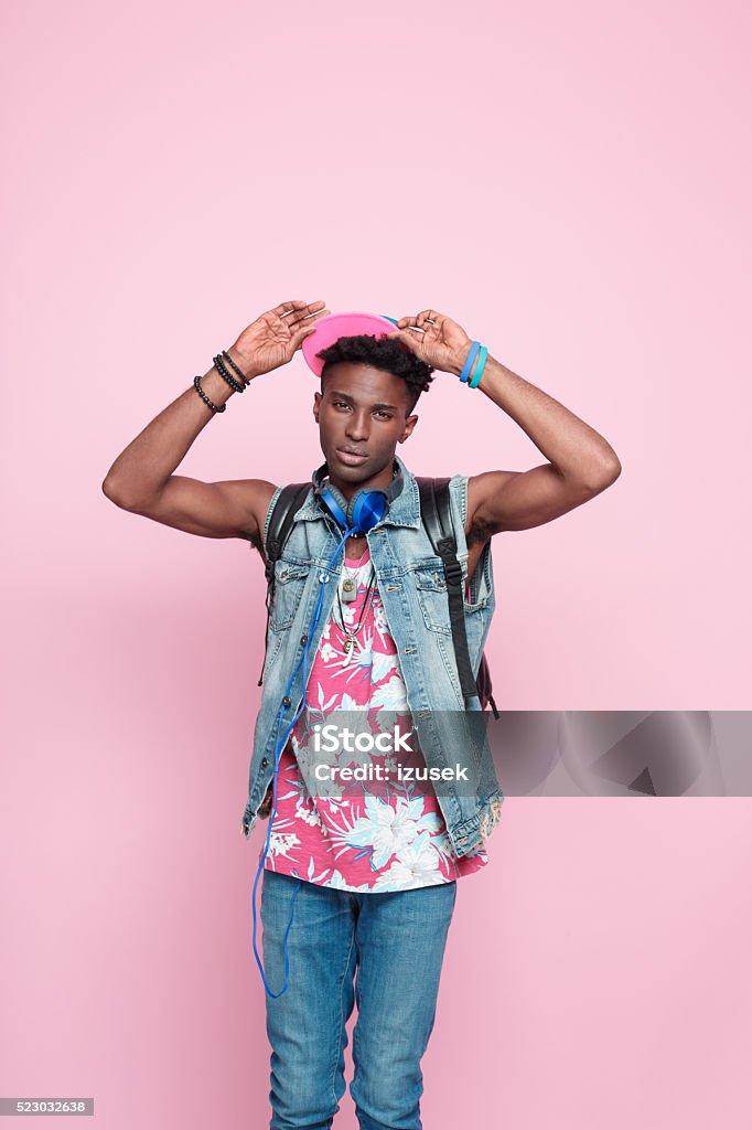 Cool afro american guy, summer studio portrait, pink background Summer portrait of carefree afro american young man wearing headphone, cap and jeans sleeveless jacket, standing against pink background, raising hands, looking at camera. Adult Stock Photo