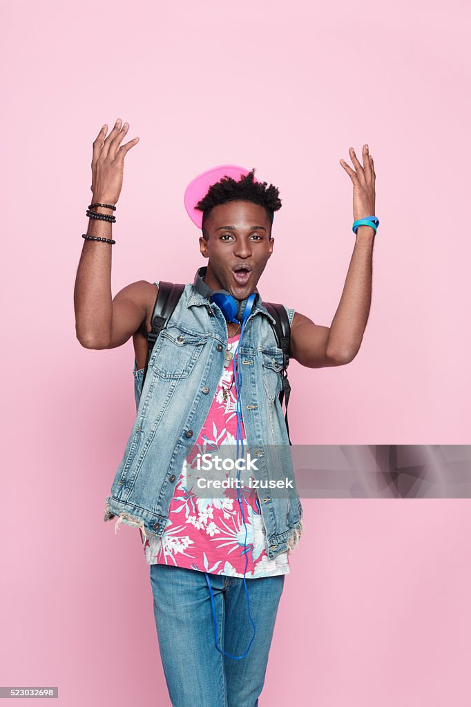Cool afro american guy, summer studio portrait, pink background Summer portrait of carefree afro american young man wearing headphone, cap and jeans sleeveless jacket, standing against pink background, raising hands, shouting. Men Stock Photo