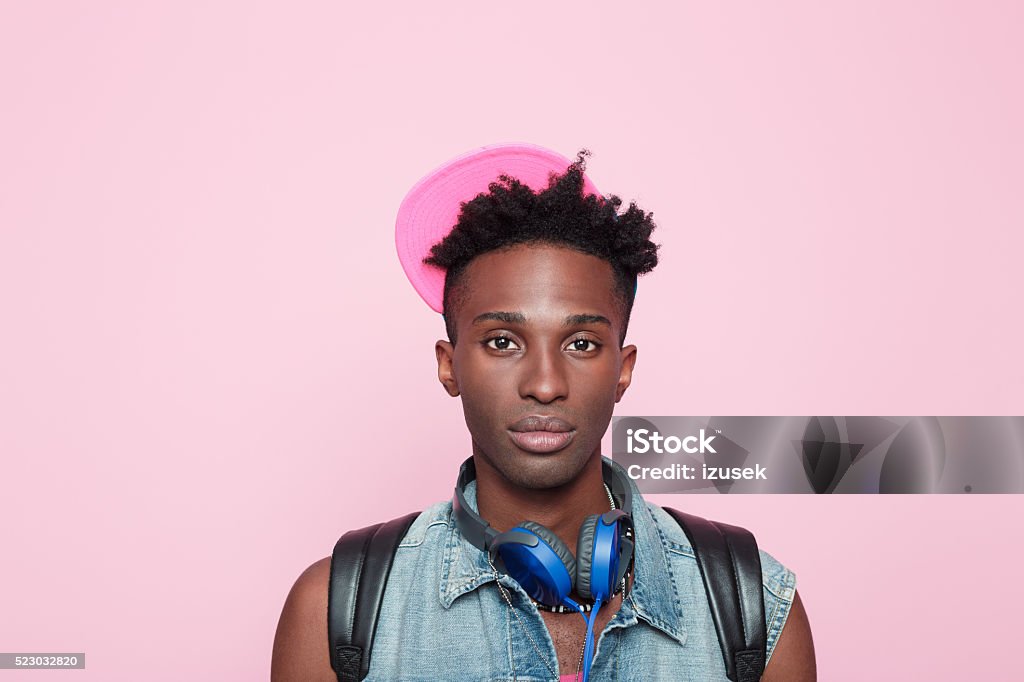 Portrait of afro american guy Portrait of sad afro american young man wearing headphone, cap and jeans sleeveless jacket, standing against pink background, looking at camera. Close up of face. Adult Stock Photo