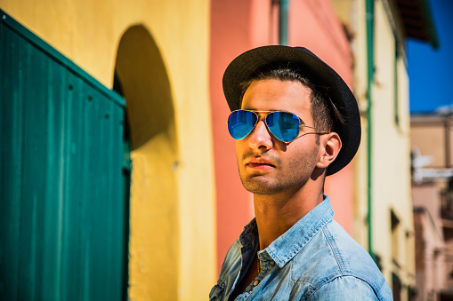 Portrait of dark haired young man in blue mirrored sunglasses and black hat. Cityscape on background, with colorful houses in Italy 