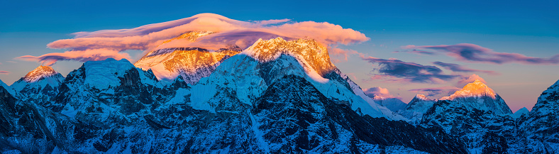 Lenticular clouds caressing the iconic pyramid peak of Mt. Everest (8848m) as golden sunset light illuminates the south west face and the summits of Nuptse (7861m), Lhotse (8516m) and Makalu (8463m) in this vibrant Himalaya mountain panorama deep in the Sagarmatha National Park of Nepal, a UNESCO World Heritage Site. ProPhoto RGB profile for maximum color fidelity and gamut.