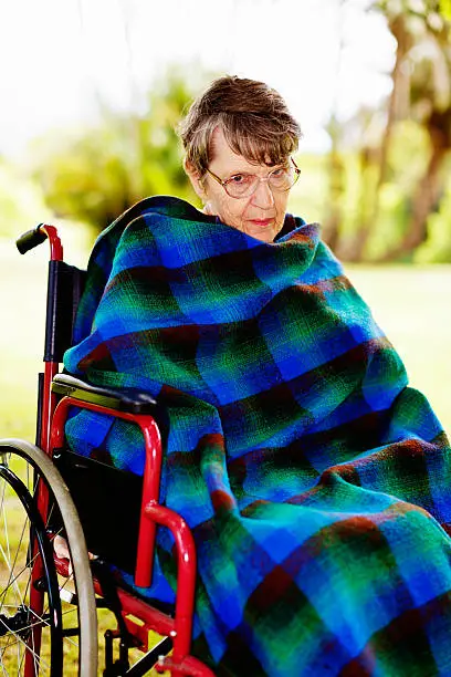 An old woman sitting in a wheelchair, outside, wrapped in a tartan blanket, looks up suspiciously. Possibly senile dementia patient.