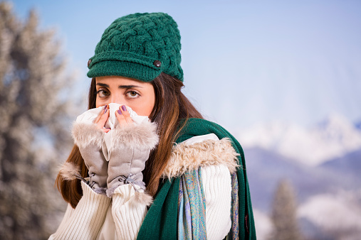 A sick, Latin descent young woman sneezes into a tissue while outside in winter.  She has the flu or allergies in winter season. 