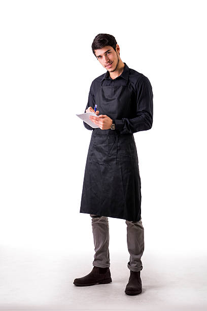 Full length shot of young chef or waiter posing isolated Full length shot of young chef or waiter posing, wearing black apron and shirt, writing order on notepad, isolated on white background electronic organizer photos stock pictures, royalty-free photos & images