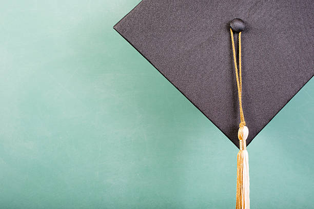Spring graduation time.  A black cap/motarboard with gold and white tassel hangs in front of a green chalkboard in classroom.  Copyspace.   No people.  Education themes.