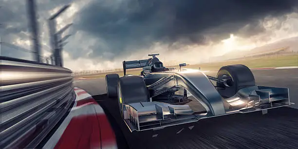 A close up image of a generic unmarked silver racing car moving at high speed around a corner close to safety barrier. The action takes place on a fictional racetrack close to an arena full of spectators under a dramatic stormy evening sky at sunset. Location is fake – mixture of CG and photographic elements. 