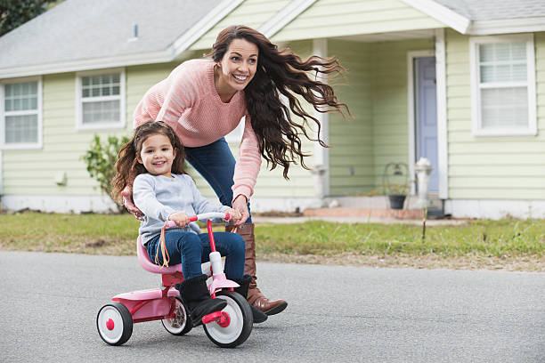 Hispanic mother helping daughter ride tricycle An Hispanic woman helping her 3 year old daughter ride a tricycle. The little girl is having a good time, smiling and looking at the camera. tricycle stock pictures, royalty-free photos & images