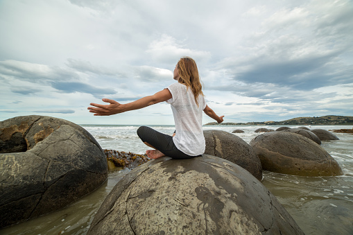 Cheering young woman sits on a boulder at Moeraki boulders, arms outstretched for freedom, positive emotions.